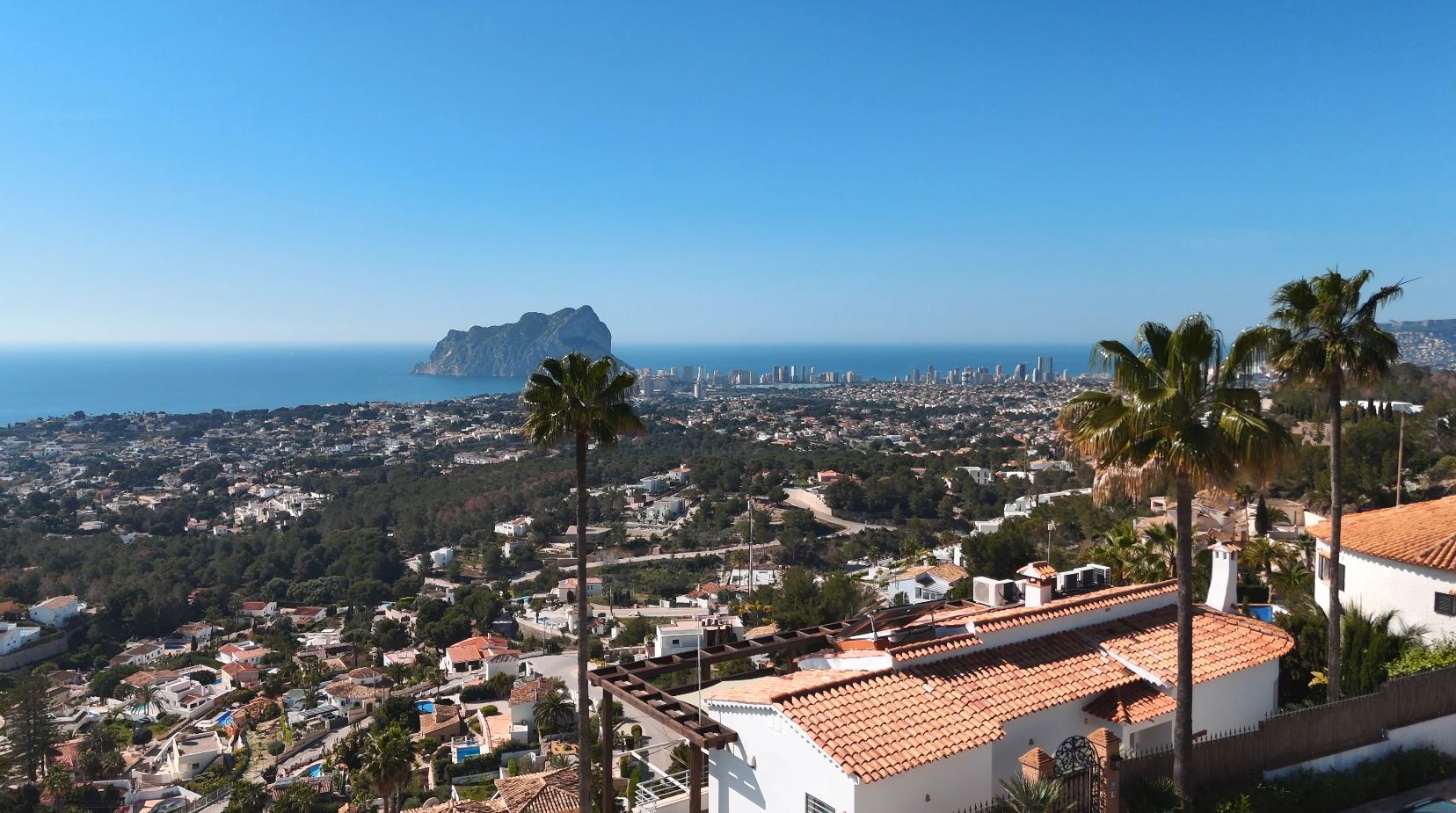 Elevated panoramic view of the Peñón de Ifach and Calpe skyline, featuring Mediterranean villas with terracotta roofs and palm trees in the foreground.