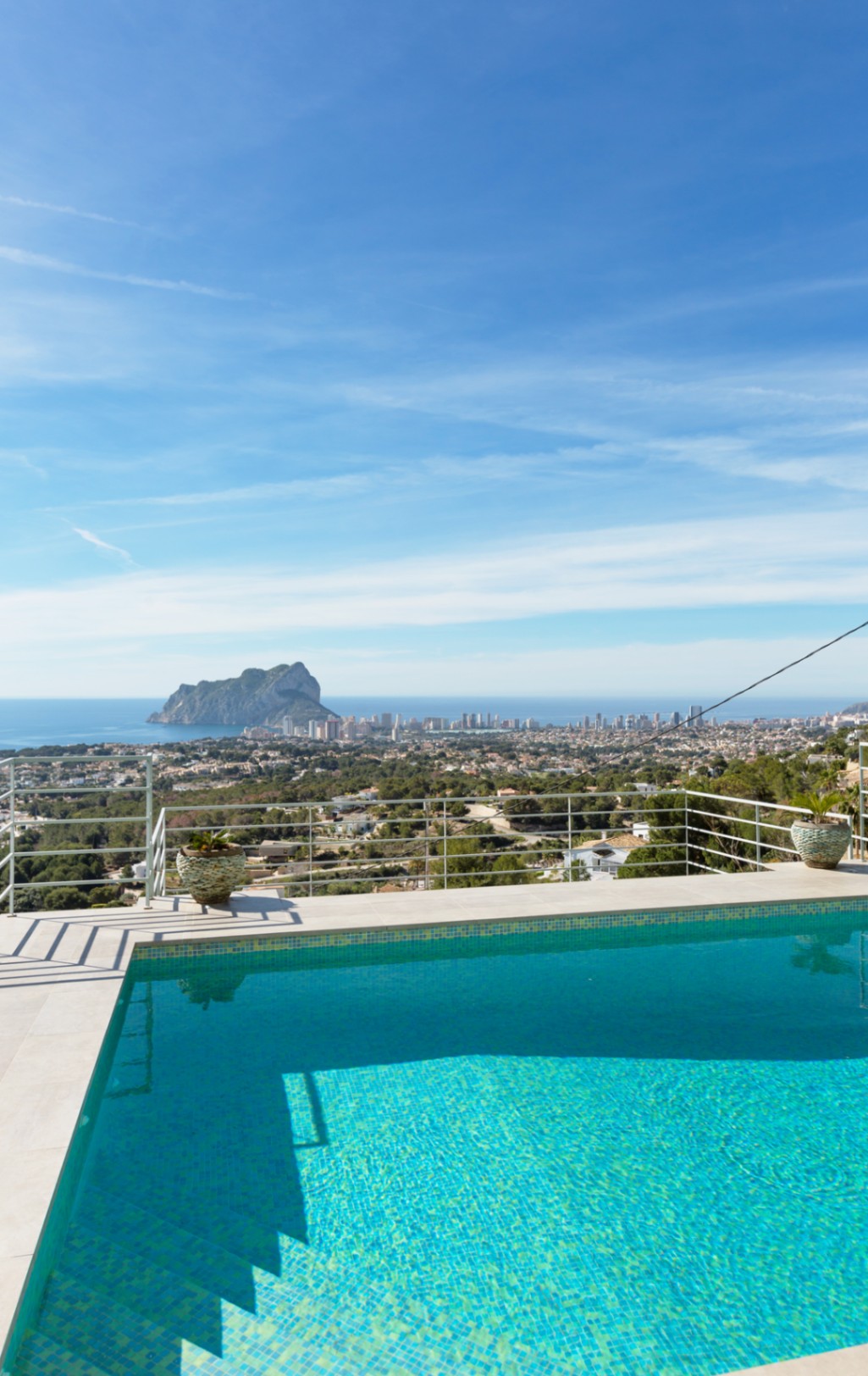 Elevated terrace featuring a turquoise mosaic-tiled pool with white stone coping, offering panoramic views of the Peñón de Ifach and the Mediterranean coastline.