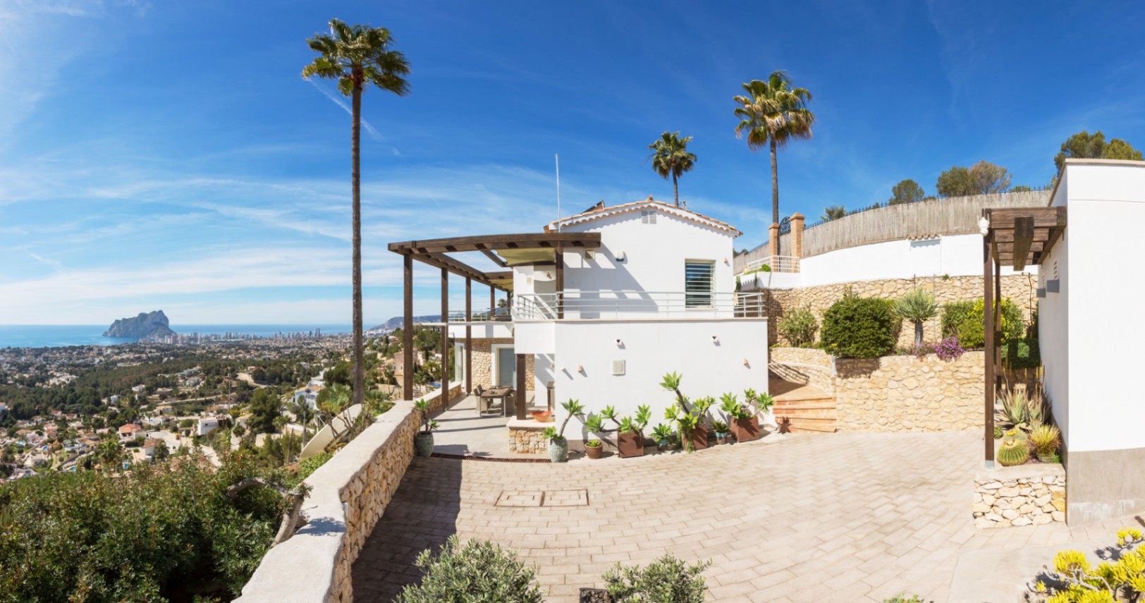 Exterior view of a Mediterranean villa with white facades, wooden pergolas, and stone walls, offering panoramic views of the coast and the Peñón de Ifach.