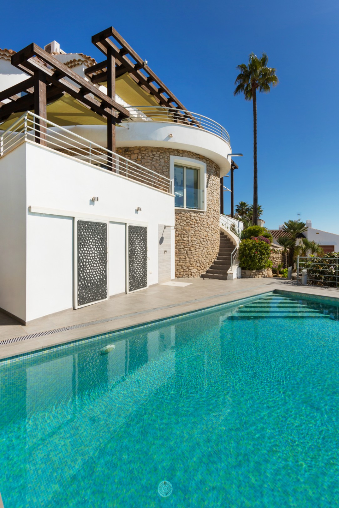 Exterior view of a turquoise tiled pool adjacent to a white villa featuring a stone-clad circular wall, wooden pergola, and decorative metal sliding panels.