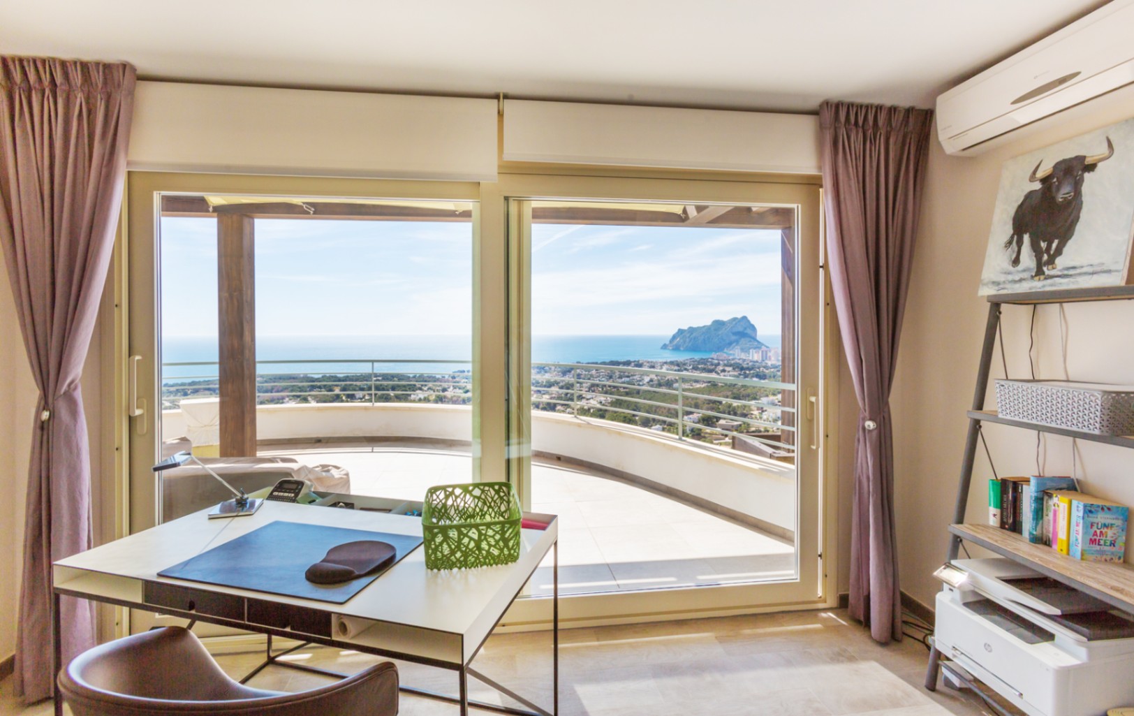 Home office with modern desk and shelving, featuring large sliding glass doors that open to a curved terrace with views of the Mediterranean and Peñón de Ifach.
