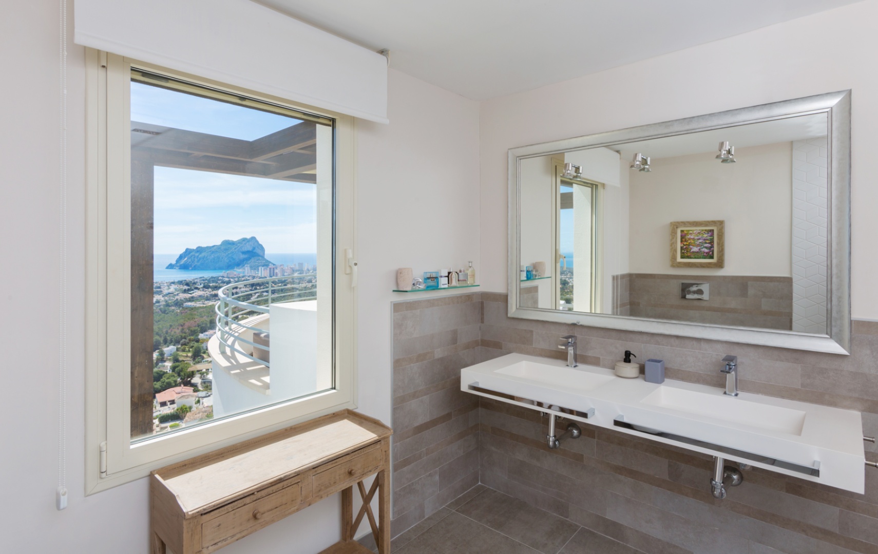 Modern bathroom featuring a double floating vanity, large silver-framed mirror, and a window offering panoramic views of the Peñón de Ifach and the coast.