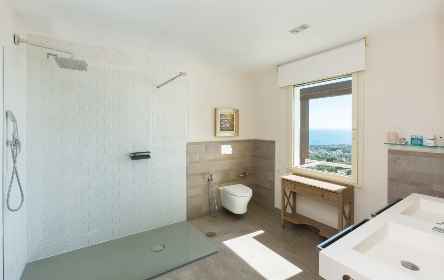 Modern bathroom featuring a walk-in shower with hexagonal tiles, a double vanity, and a large window offering panoramic sea views over the coastline.