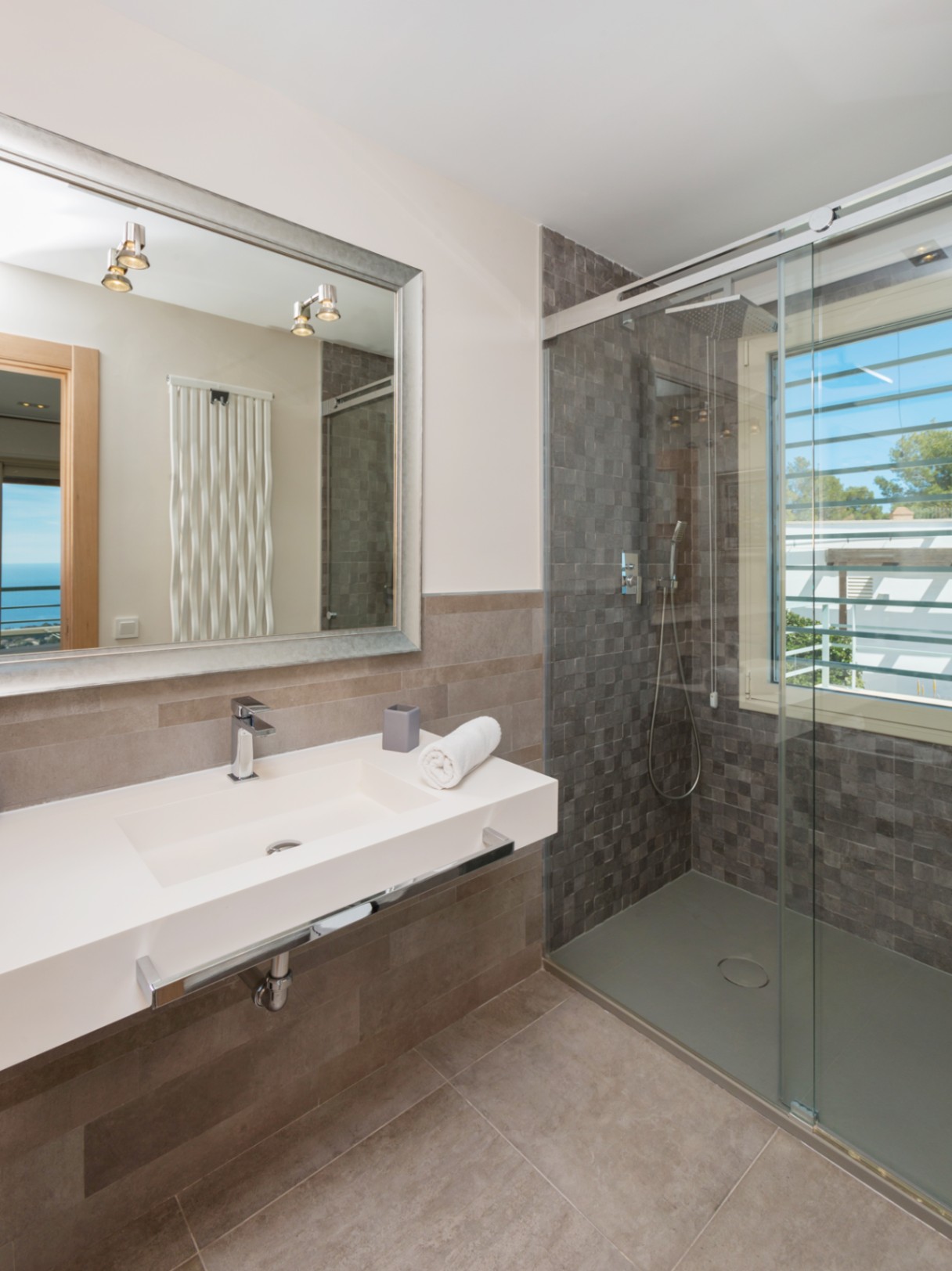 Modern bathroom featuring a white integrated sink, a large silver-framed mirror, and a walk-in shower with grey mosaic tiles and natural light.