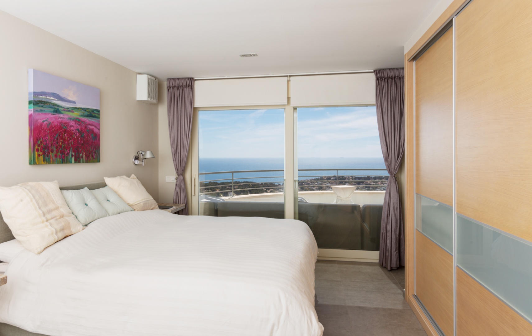 Modern bedroom featuring a large bed, built-in wooden wardrobe, and floor-to-ceiling glass doors opening to a terrace with expansive sea views.