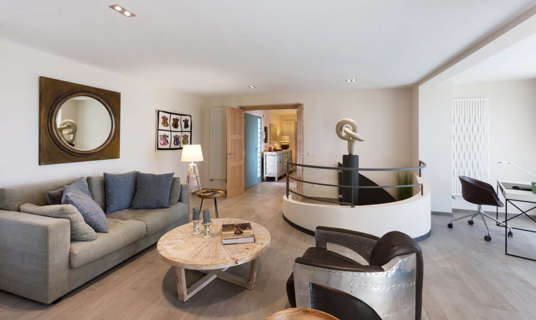 Modern living area featuring a grey sofa, aviator-style armchair, and circular staircase with a central sculpture. Includes a workspace and light wood flooring.