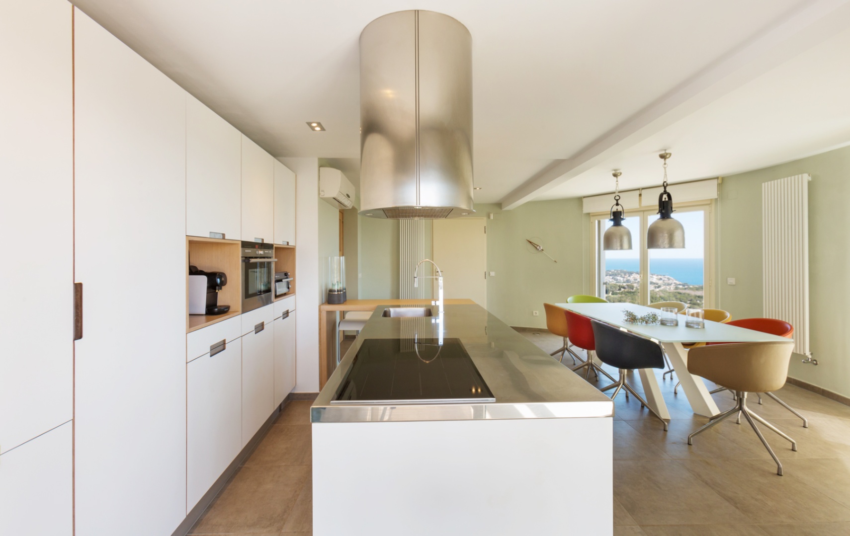 Modern open-plan kitchen featuring a white island with a stainless steel top, cylindrical extractor, and a dining area with sea views through glass doors.