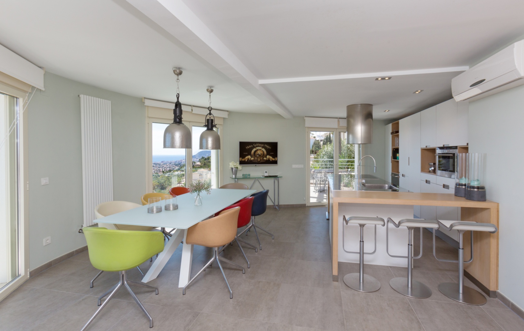 Open-plan interior featuring a modern kitchen with a wooden breakfast bar and a dining area with colorful chairs and industrial lighting overlooking the coast.