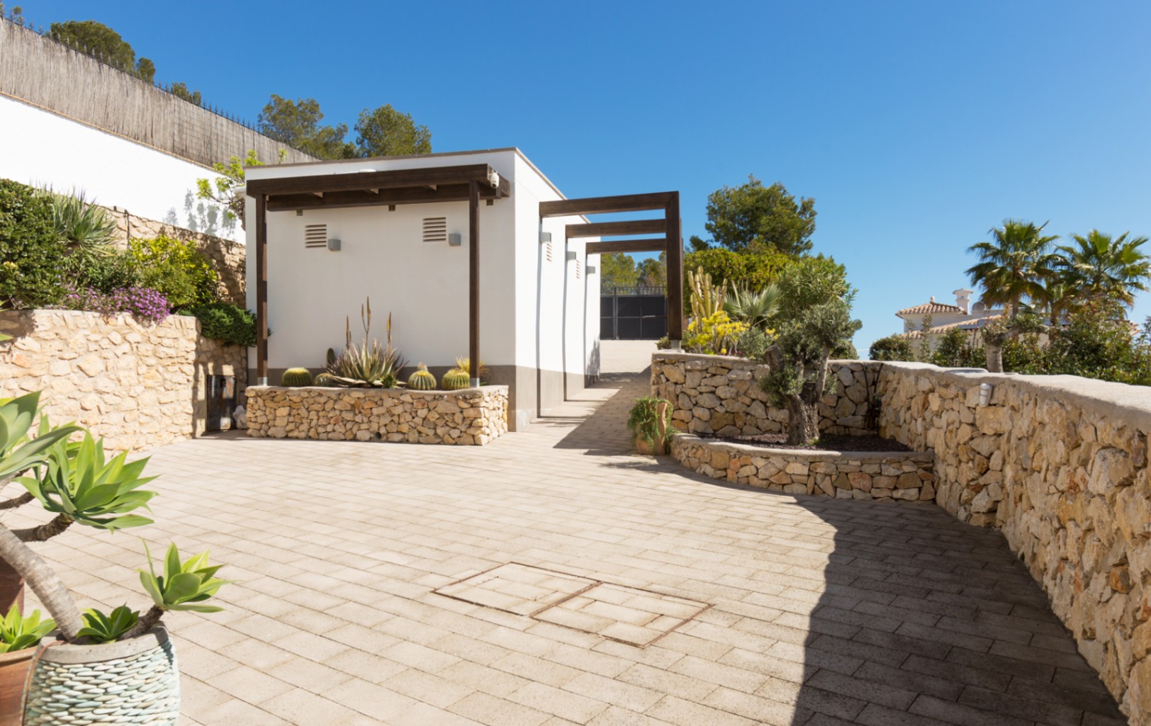 Paved outdoor area featuring dry stone walls, raised planters with cacti, and a white modern structure with dark wooden beams.