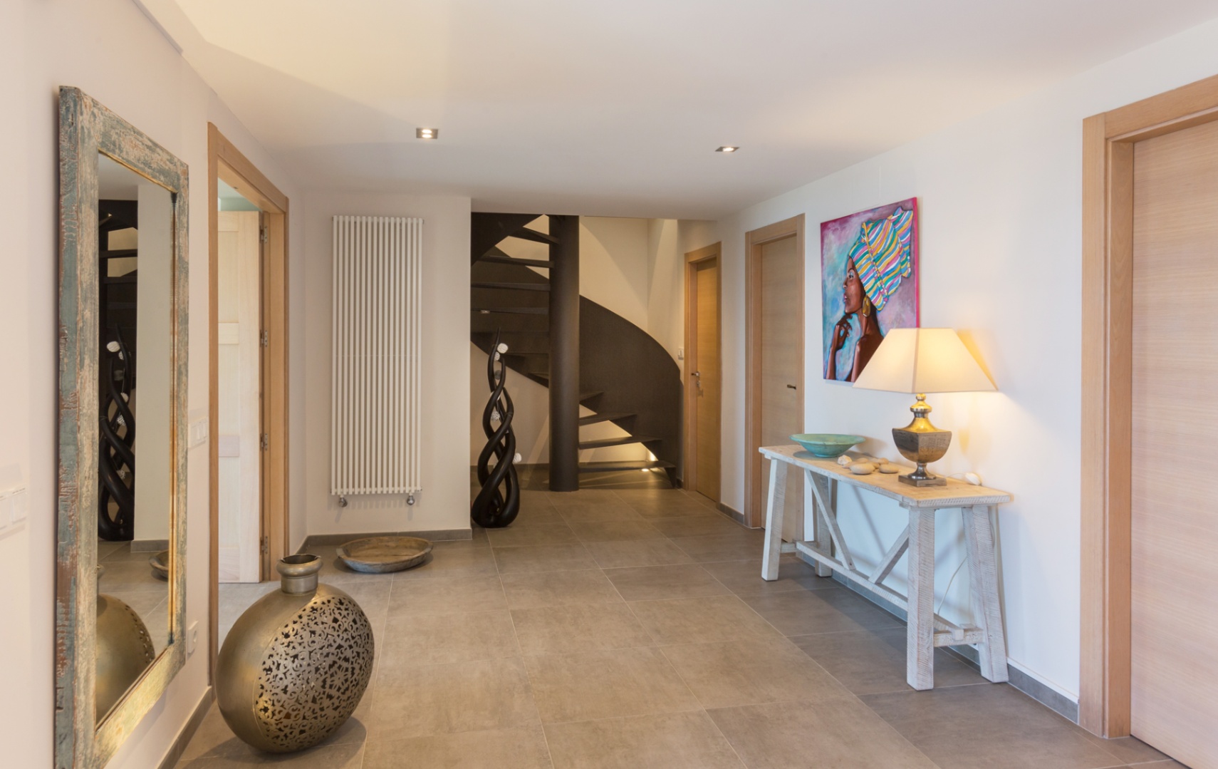 Spacious hallway with grey tiled floors, featuring a dark metal spiral staircase, light wood doors, and a rustic console table with a decorative lamp and art.