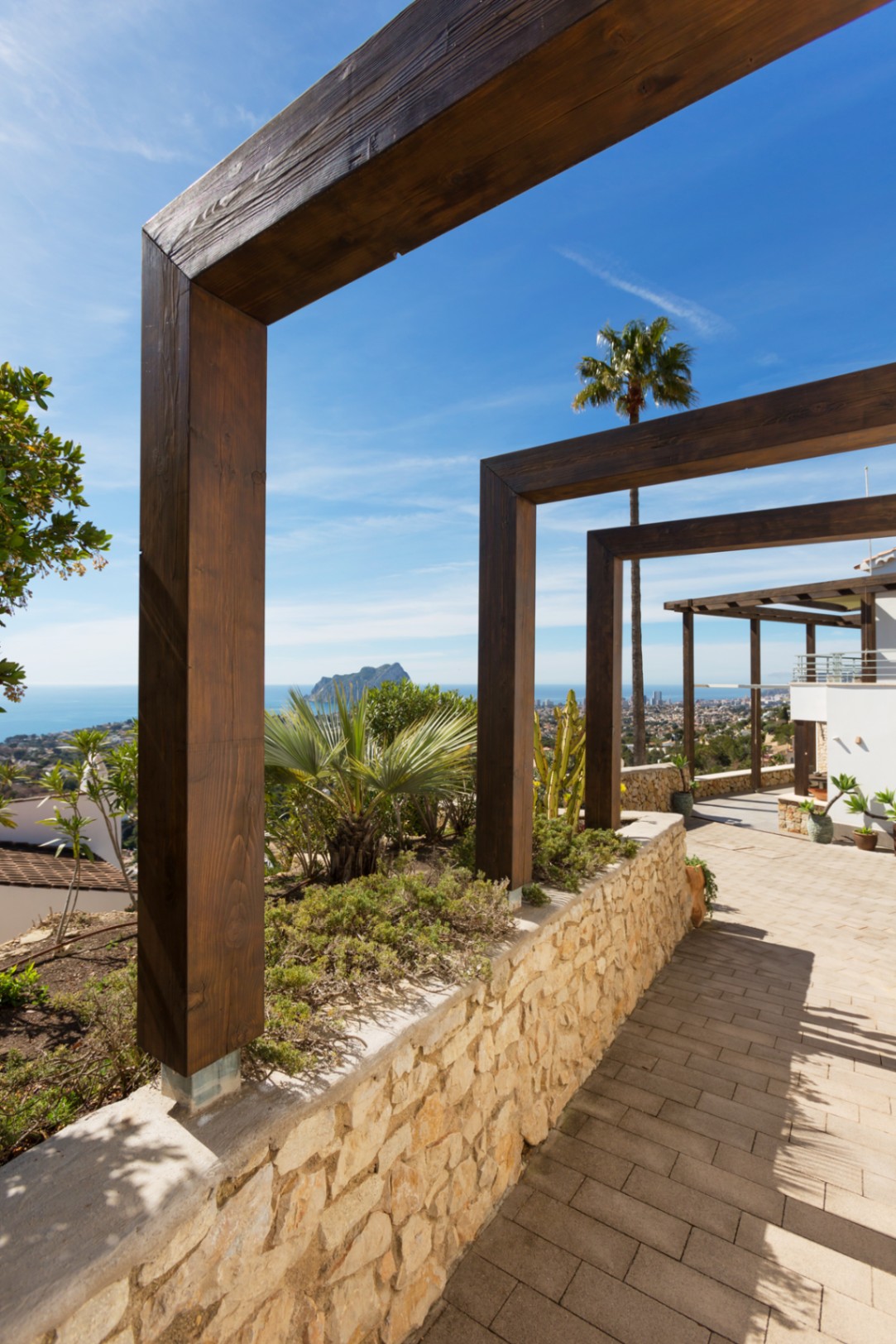 Terrace walkway featuring dark wood pergola frames, a natural stone wall, and panoramic Mediterranean views including the Peñón de Ifach.