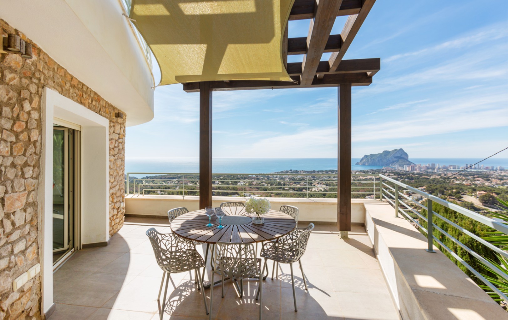 Terrace with natural stone walls, a dark wood pergola, and a sail shade. Features a round wooden dining table with panoramic views of the Mediterranean and Peñón de Ifach.