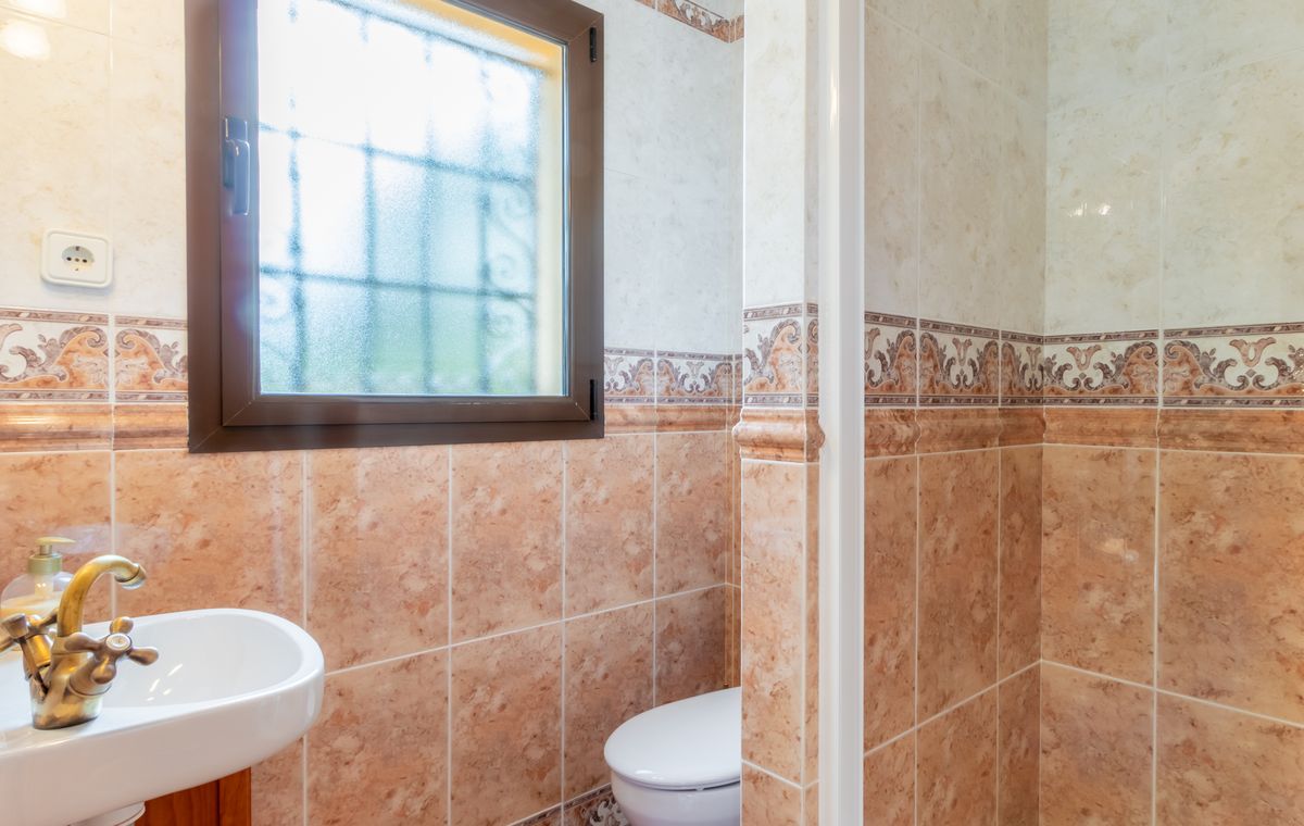 Bathroom featuring marble-effect terracotta tiles, a vintage brass faucet on a white sink, and a frosted window with security bars for privacy.