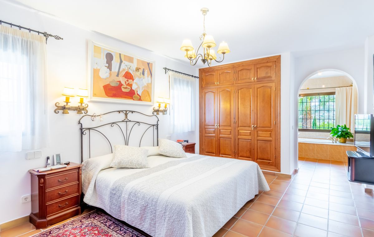 Bedroom featuring a wrought iron headboard, built-in wooden wardrobes, and terracotta flooring. An arched doorway leads to an ensuite bathroom with a built-in tub.