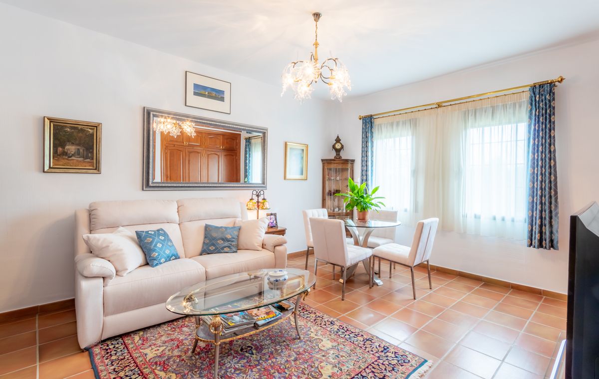 Bright living area featuring terracotta tile flooring, a white sofa with blue accents, and a glass dining set under an ornate glass chandelier.
