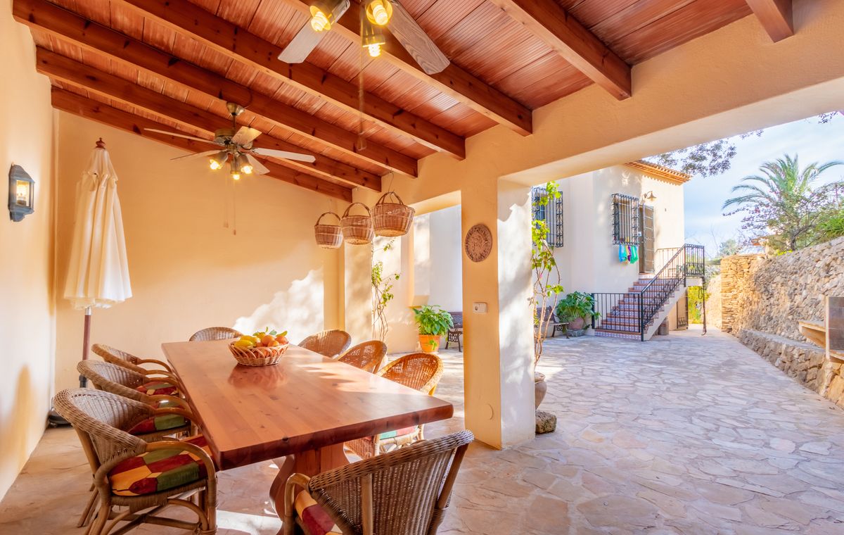 Covered terrace featuring a wooden beamed ceiling with fans, a large dining table, and wicker seating overlooking a stone-paved courtyard.