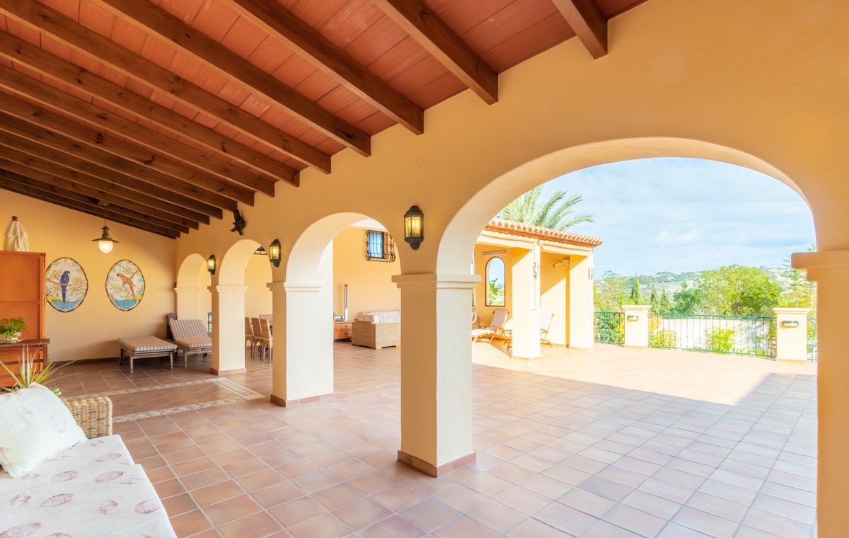 Covered terrace featuring dark wood beams, terracotta tile flooring with decorative inlays, and arched columns overlooking an expansive open patio.
