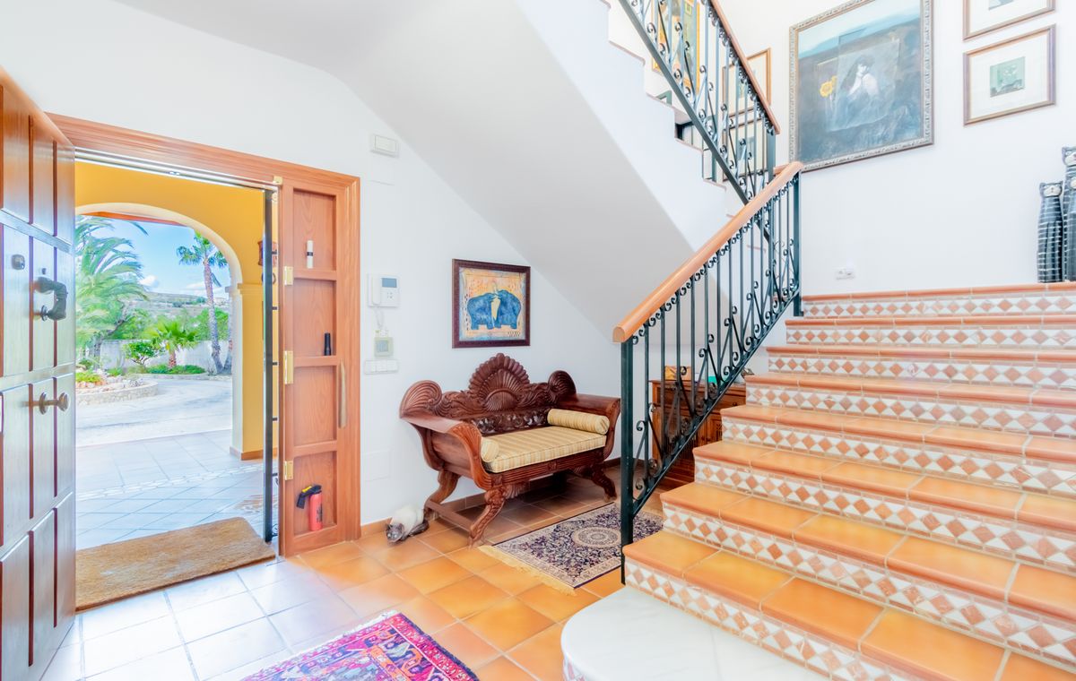 Entry hall featuring a terracotta-tiled staircase with decorative risers, wrought iron railing, and an ornate carved wooden bench. The open front door reveals a sunny patio.