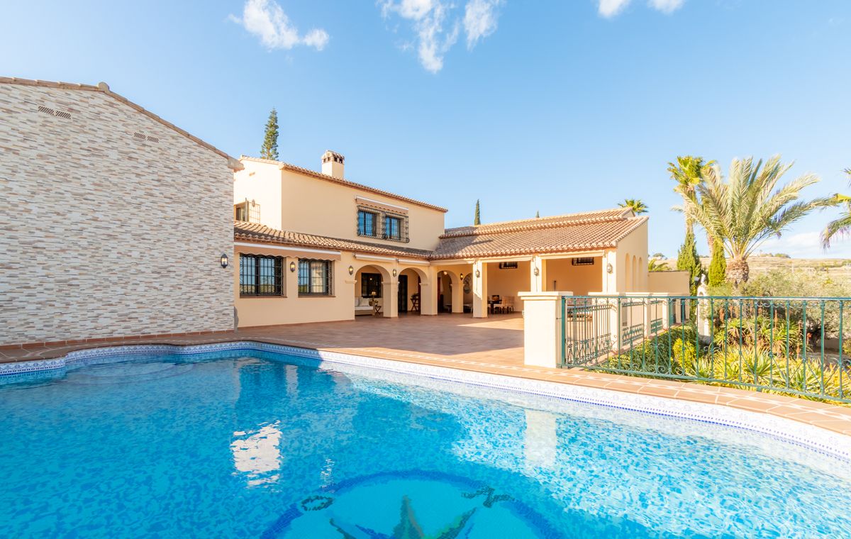 Exterior view of a Mediterranean villa featuring a large swimming pool with mosaic detailing, a terracotta terrace, and a covered arched porch.