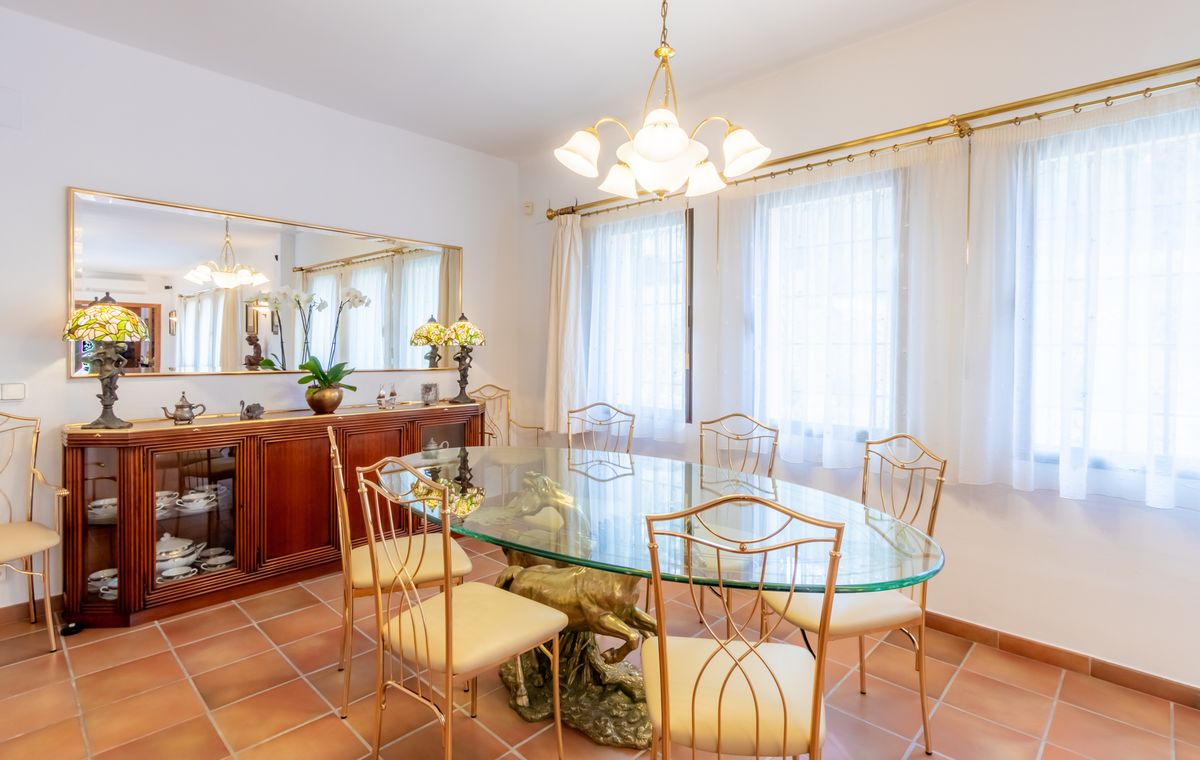 Formal dining room featuring an oval glass table with a sculptural brass base, gold-toned chairs, terracotta flooring, and a large wooden sideboard.
