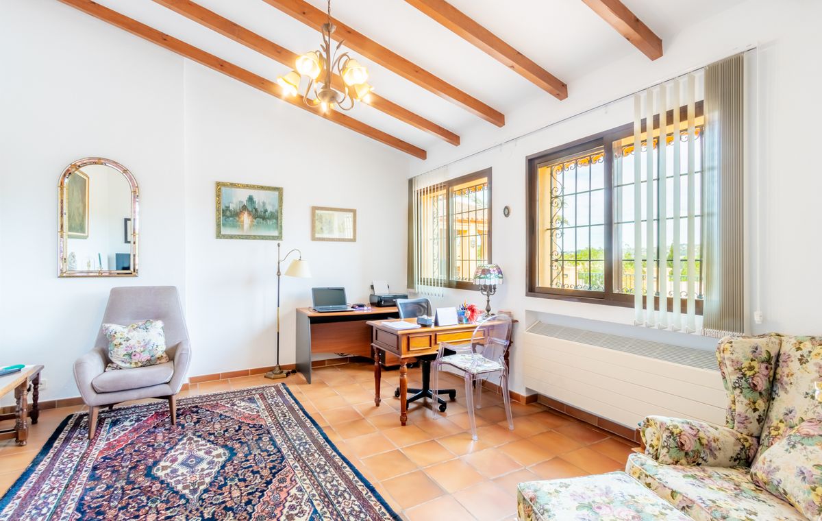 Home office featuring exposed wooden ceiling beams, terracotta tile flooring, and dual workspaces. Large windows with security grilles provide natural light.