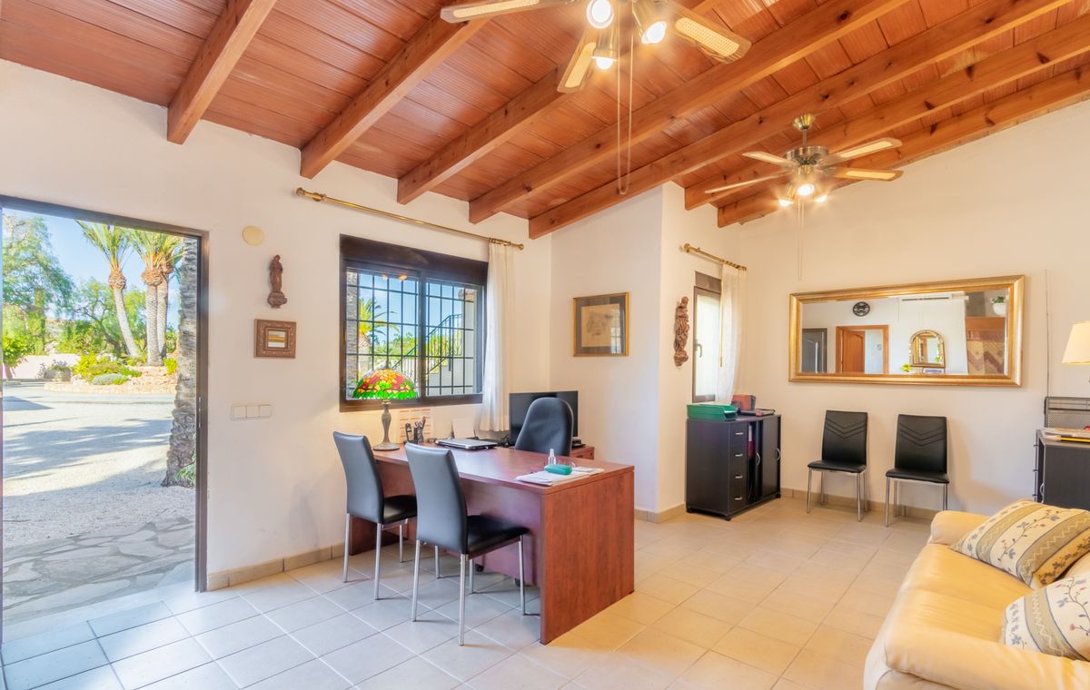 Home office with exposed wooden beam ceilings, ceiling fans, and a wooden desk. An open door reveals a garden view with palm trees and natural light.