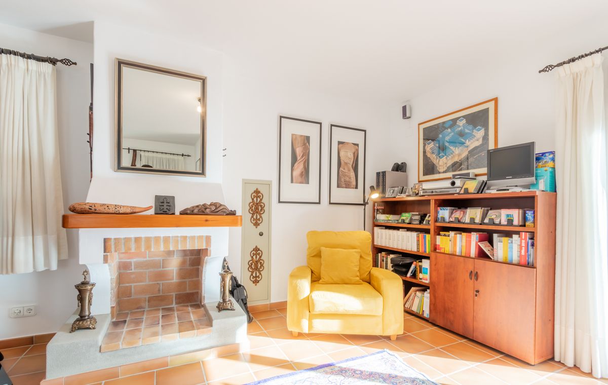 Living area featuring a traditional brick fireplace, a yellow armchair, and a large wooden bookshelf on terracotta tile flooring.