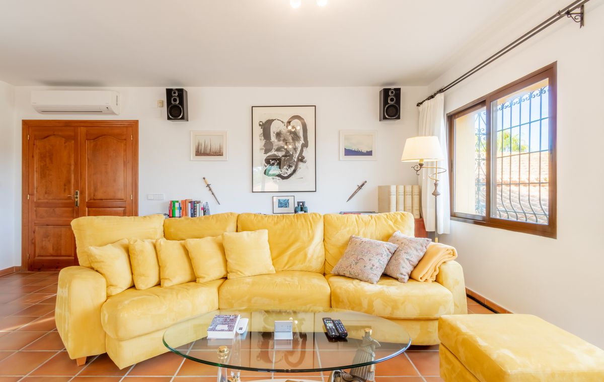 Living room featuring a large yellow corner sofa, glass coffee table, and terracotta tile flooring. The white walls are decorated with artwork and integrated speakers.