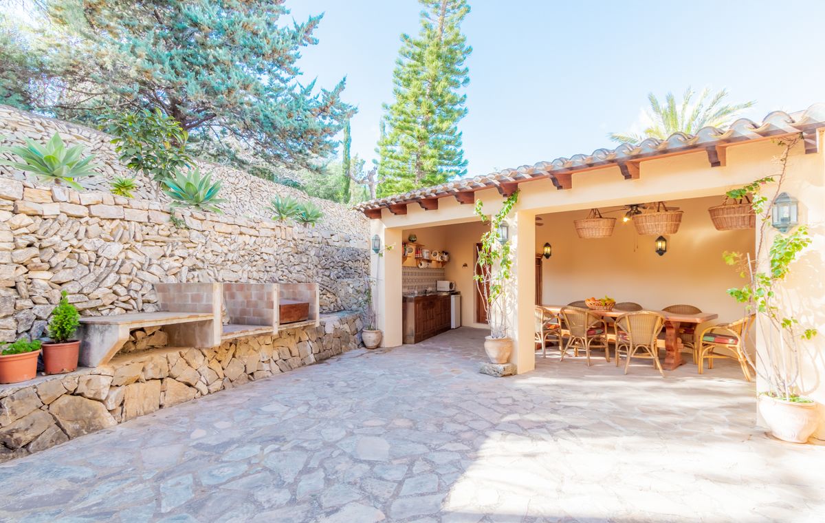 Outdoor dining area featuring a stone-paved patio, built-in barbecue, and a covered porch with exposed beams, wicker seating, and a functional kitchenette.