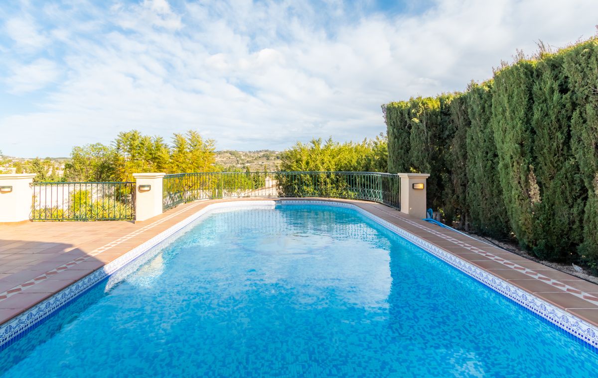 Outdoor swimming pool with blue mosaic tiling and terracotta borders, featuring a black metal safety railing and a tall cypress hedge for privacy.