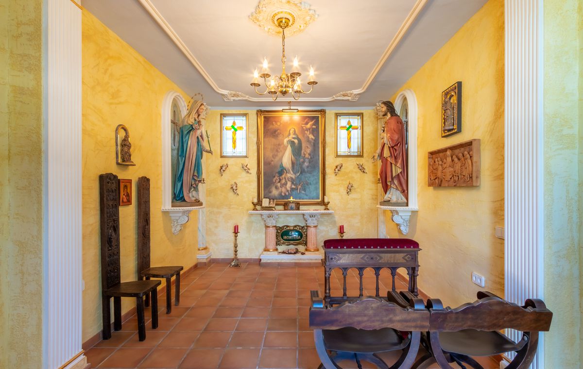 Private chapel featuring yellow textured walls, terracotta flooring, a central altar with religious paintings, statues in niches, and traditional wooden seating.