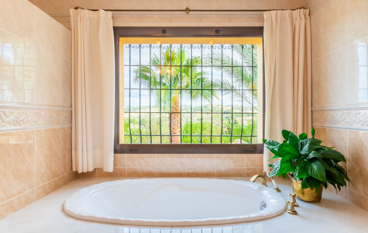 Recessed oval bathtub with brass fixtures, positioned beneath a large window with wrought iron bars and views of palm trees and the countryside.