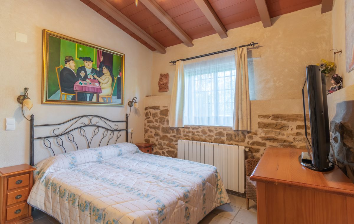 Rustic bedroom featuring a wrought iron bed, exposed stone walls, and a beamed wooden ceiling. A large figurative painting and a wooden dresser with TV complete the space.