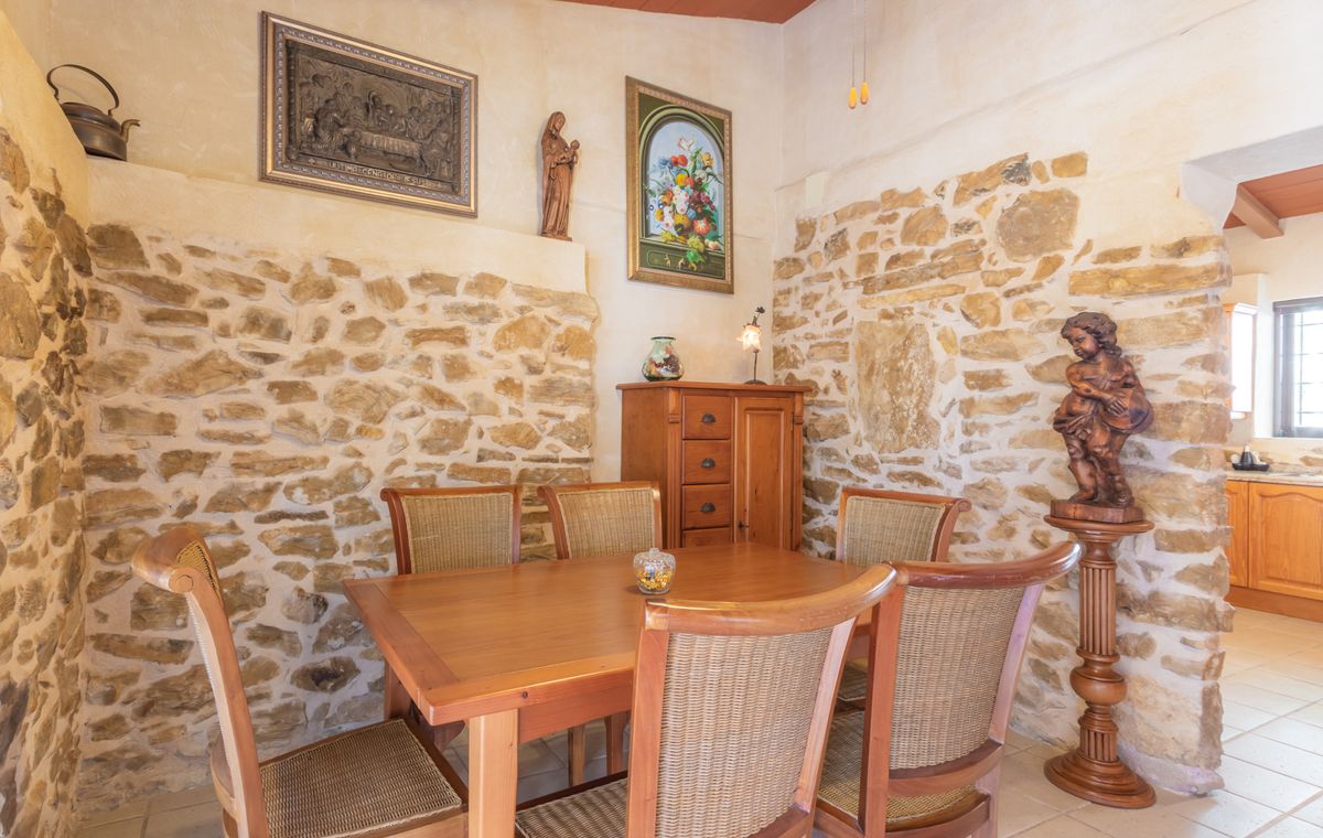 Rustic dining area featuring exposed natural stone walls, a solid wood table with wicker-back chairs, and traditional religious artwork and sculptures.