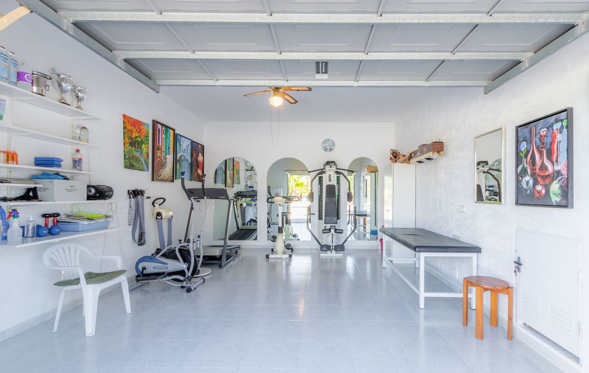 Spacious home gym in a converted garage with tiled floors, featuring cardio machines, a multi-gym station, and a treatment table under a ceiling fan.