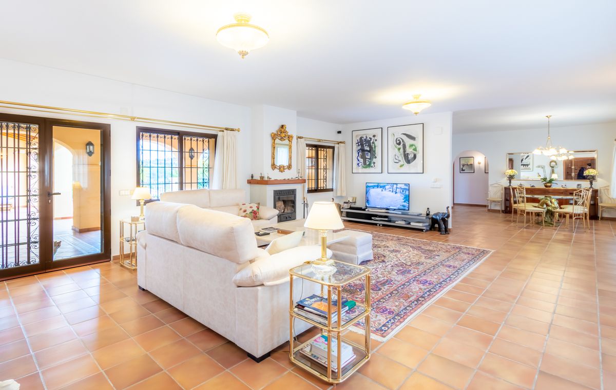 Spacious living area with terracotta flooring, featuring a cream sectional sofa, a fireplace with a gold-framed mirror, and a connected dining space.