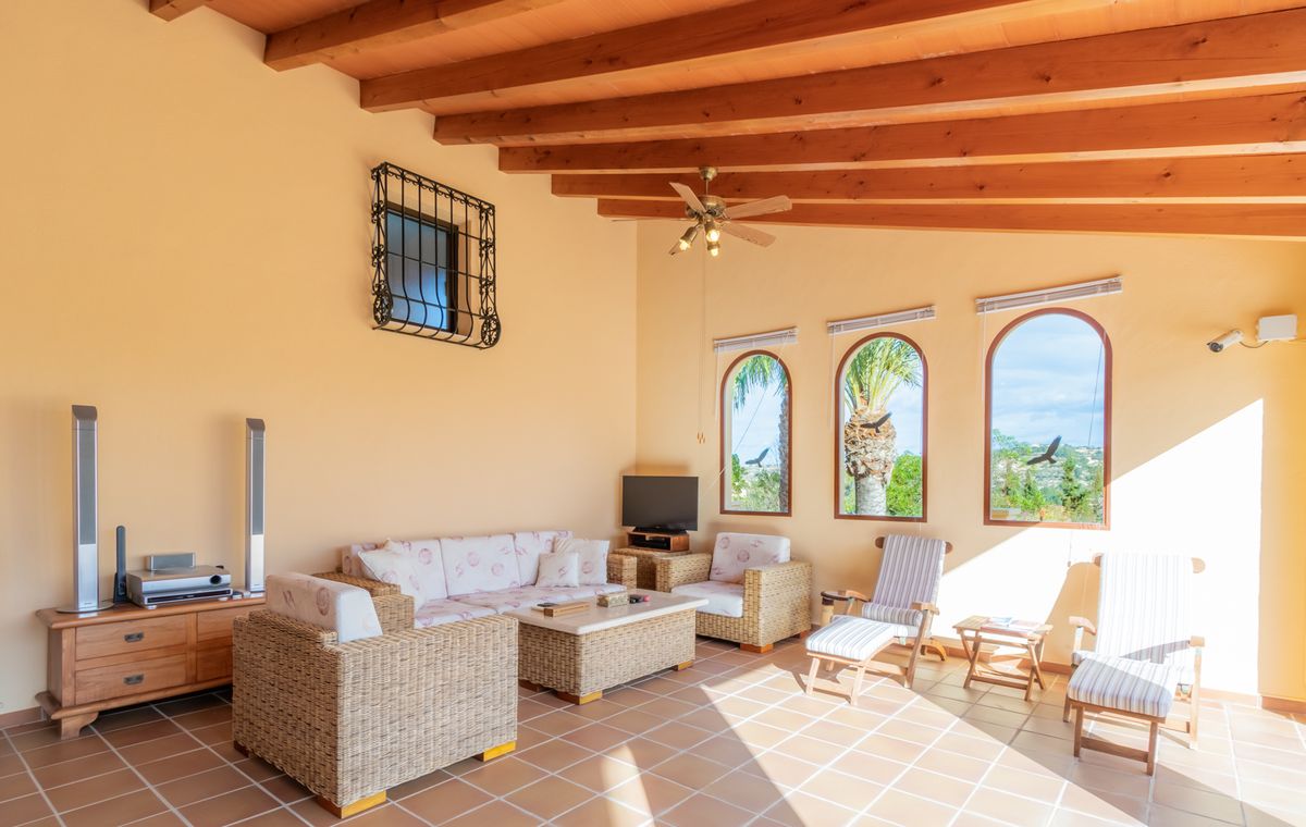 Sunroom featuring exposed wooden beams, terracotta flooring, and wicker furniture. Three arched windows provide natural light and views of the surrounding palm trees.