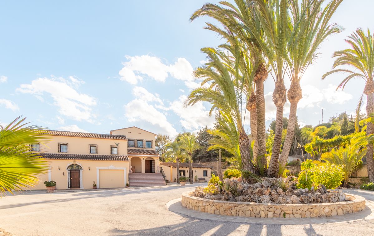 Wide paved entrance driveway featuring a circular stone planter with palm trees. The villa displays cream-colored facades and traditional terracotta roof tiles.