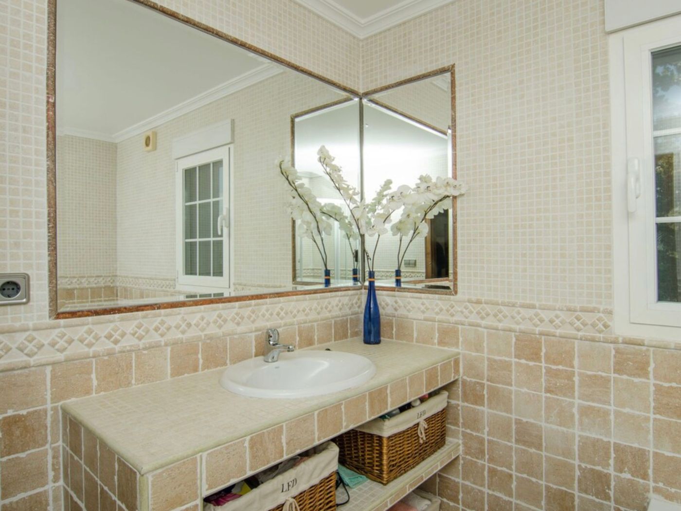 Bathroom featuring a tiled vanity with an integrated sink and open shelving with wicker baskets. Large corner mirrors reflect the neutral mosaic tile walls.