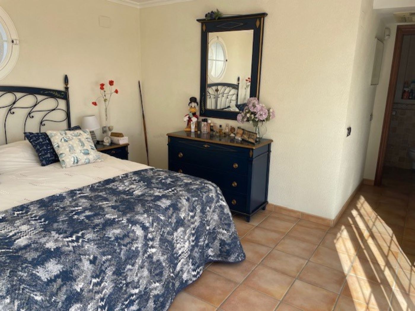 Bedroom featuring a wrought iron bed frame, a navy blue dresser with a matching mirror, and traditional terracotta tile flooring under natural light.