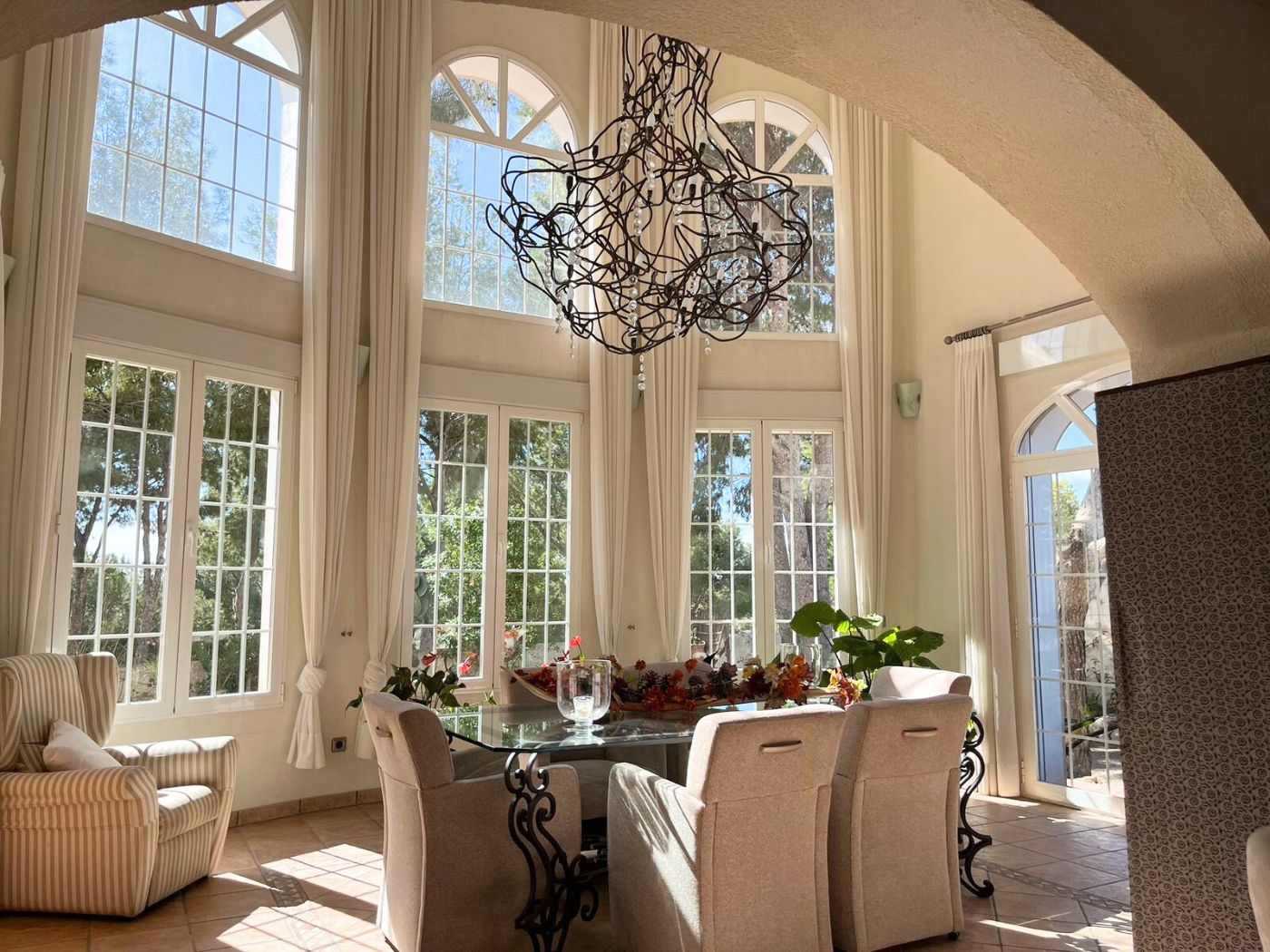 Bright dining area featuring double-height ceilings, arched windows, a glass-top table with wrought iron legs, and a sculptural wire chandelier.
