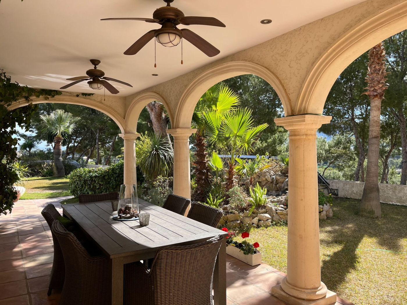 Covered terrace featuring a stone arched colonnade, terracotta flooring, and a large dining set overlooking a lush Mediterranean garden with palm trees.