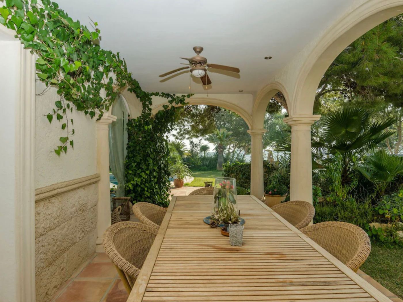 Covered terrace featuring stone arches, a long wooden dining table with wicker chairs, and a ceiling fan, overlooking a lush Mediterranean garden.