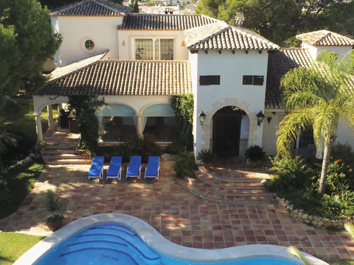 Elevated view of a Mediterranean villa featuring a curved pool, terracotta terrace with four sun loungers, and a white facade with arched stone entrances.
