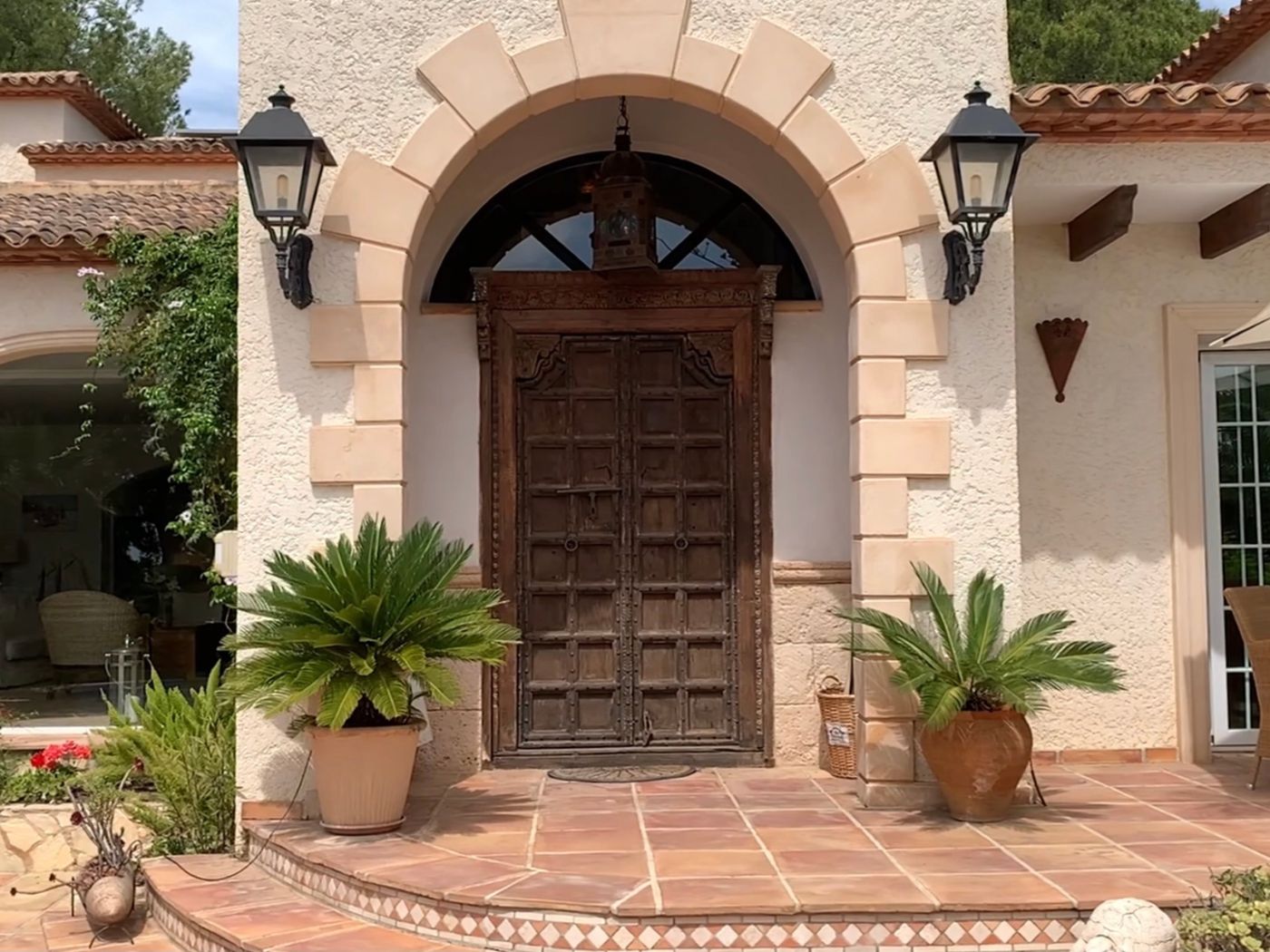 Entrance featuring an antique carved wooden double door set within a stone arch, flanked by traditional black lanterns and potted sago palms on a terracotta porch.
