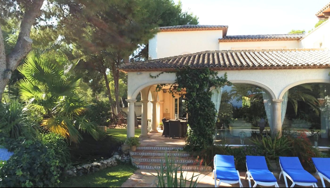 Exterior of a Mediterranean villa featuring an arched porch with stone columns, a terracotta roof, and a terrace with blue sun loungers.