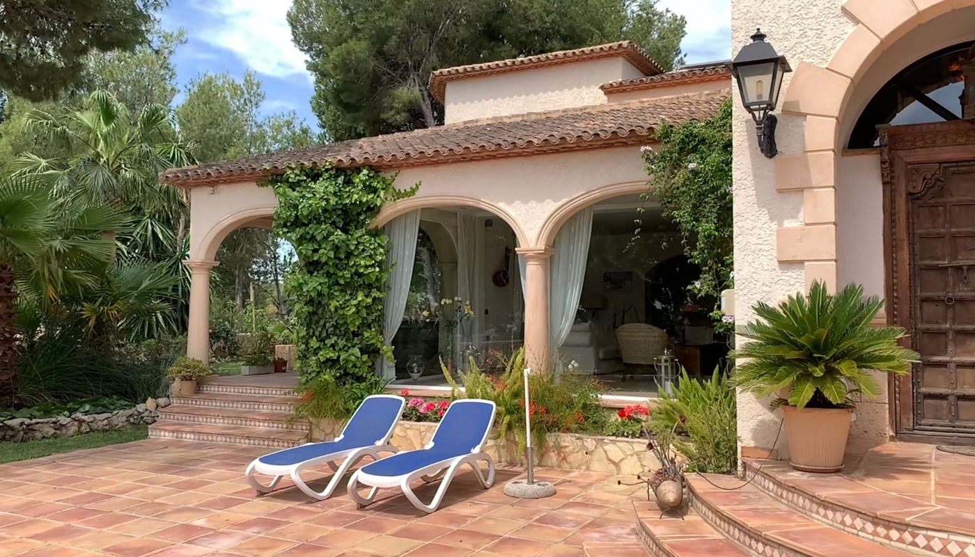 Exterior terrace featuring terracotta tiling, blue sun loungers, and an arched porch with climbing plants adjacent to a traditional carved wooden door.