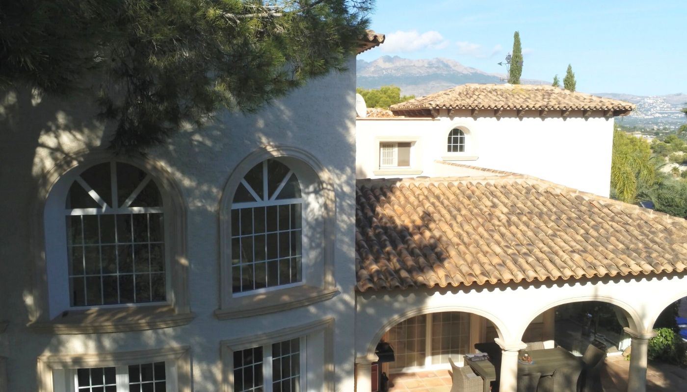 Exterior view of a Mediterranean villa featuring terracotta tiled roofs, arched windows, and a covered terrace with mountain views.