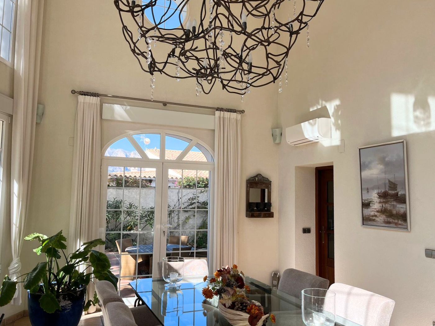 High-ceilinged dining room featuring a glass table and an ornate wrought-iron chandelier. Arched doors lead to a sunlit patio with outdoor seating.