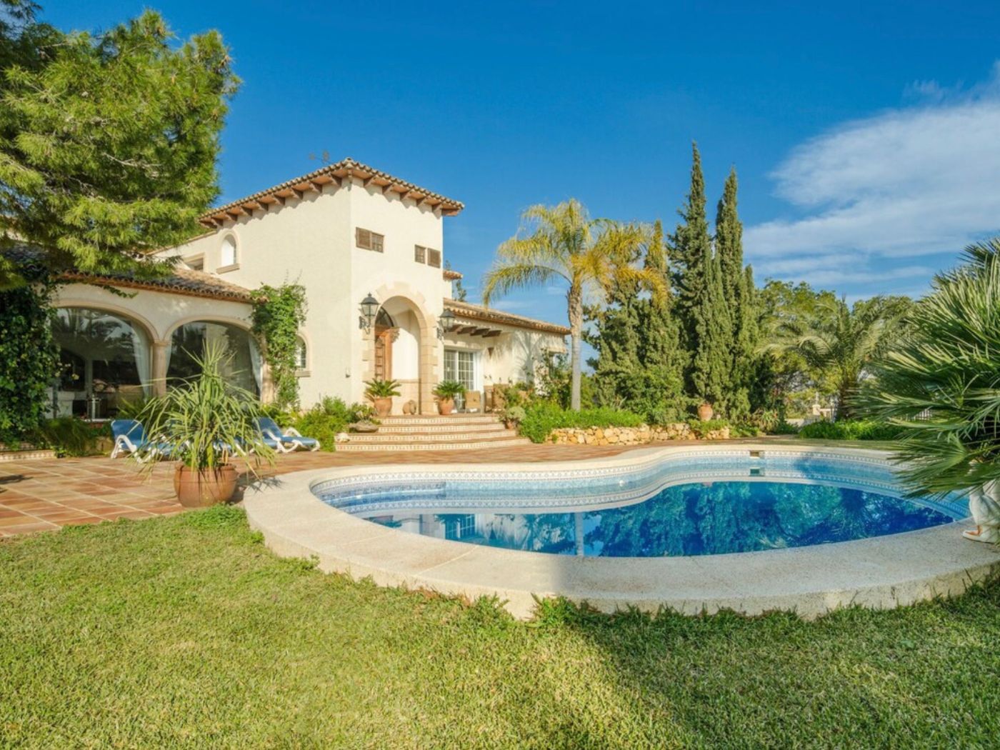 Mediterranean villa exterior featuring a curved swimming pool, terracotta patio, and white stucco facade with traditional arched details and mature landscaping.